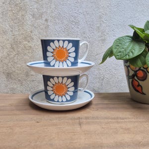 Puede incluir: Dos tazas y platillos de cerámica blanca apilados con un diseño de margarita en azul y naranja. Cada taza tiene un asa curva y el platillo presenta una banda circular azul a juego en el borde. El diseño es consistente en ambas piezas.
