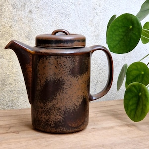 May include: A cylindrical ceramic teapot featuring a mottled dark brown and black glaze. The teapot has a flat lid with a small central handle, a wide spout, and a curved side handle. The finish displays a textured, uneven appearance.