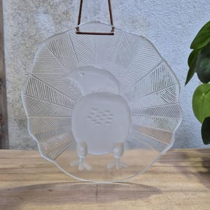 Op de afbeelding: Een rond ornament van helder glas met een mat reliëf van een vogel met een uitwaaierende staart. Het glas heeft geëtste lijndetails op de staartveren en is opgehangen aan een dun bruin leren koord dat aan de bovenrand is bevestigd.