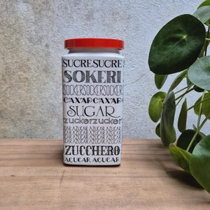 Op de afbeelding: Een witte rechthoekige voorraadbus met een rood deksel, voorzien van het woord suiker in verschillende talen en lettertypes, waaronder SUCRE, SOKERI, SOCKER, CAXAP, SUGAR, zucker, AZUCAR, ZUCCHERO en ACUCAR op de gehele voorzijde.