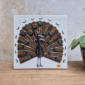 May include: A square white ceramic wall tile featuring a raised, stylized peacock with a fanned tail. The peacock is rendered in shades of brown, blue, and green, with circular accents on the feathers. A small gold sticker is in the bottom right corner.
