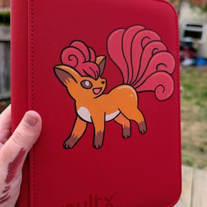 May include: A red zippered binder featuring a graphic of a Vulpix character on the front cover. The Vulpix is orange with a pinkish-red curled tail and tuft of hair. The word vaultx is embossed in the lower left corner of the red textured cover.