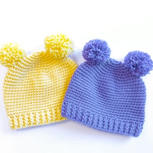 以下が含まれることがあります： ポンポンが付いた手編みの帽子が2つ。1つは黄色、もう1つは紫です。
