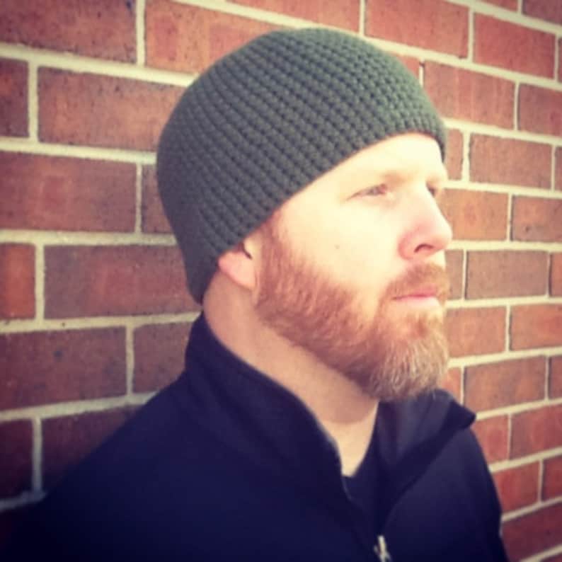 May include: A man wearing a dark green knitted beanie with a thick, textured weave. He has a full, reddish-brown beard and is looking off to the side.