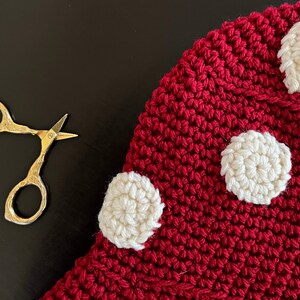 May include: A red crocheted mushroom with white crocheted polka dots. The mushroom is lying on a black surface.