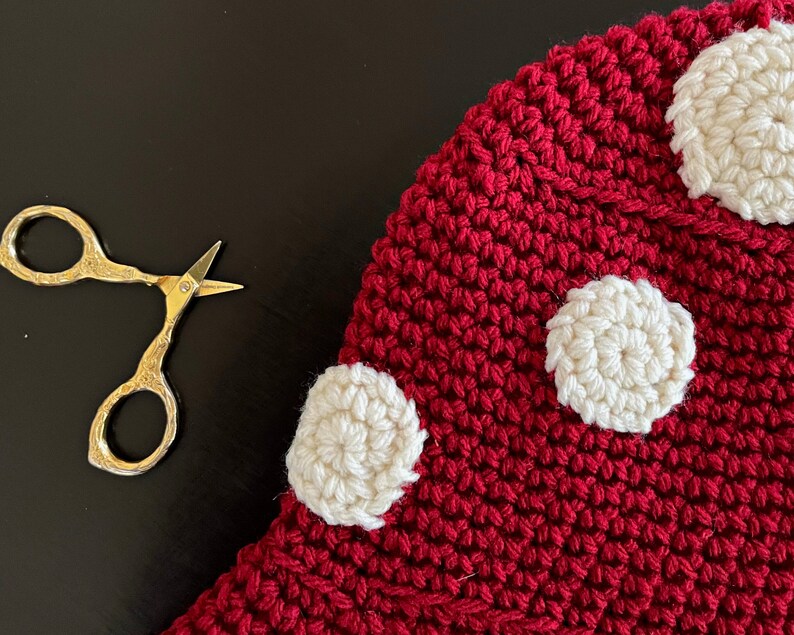 May include: A red crocheted mushroom with white crocheted polka dots. The mushroom is lying on a black surface.