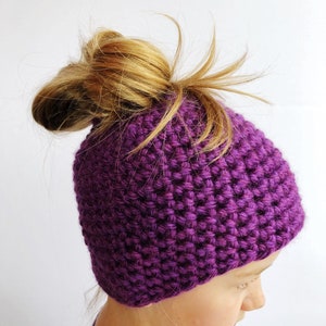 May include: Purple knitted hat with a hole in the top for a ponytail. The hat is made of a soft, textured yarn.