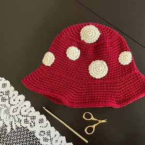 May include: A red crocheted hat with white polka dots. The hat is lying on a dark surface with a white crocheted lace doily and a gold crochet hook.
