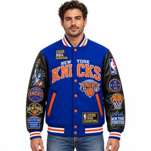 Può includere: Una giacca varsity blu con maniche nere caratterizzata dal marchio New York Knicks, con il testo 2026 NBA CHAMPIONS, la Statua della Libertà, grafiche di pallacanestro e toppe del Madison Square Garden. Presenta bordi a righe arancioni e bianchi.