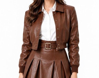 Peut inclure: Une veste en similicuir marron avec col, boutons-pression et poignets côtelés, associée à une mini-jupe plissée taille haute assortie dotée d'une ceinture à boucle dorée, portée sur une chemise blanche à col boutonné.