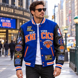 Peut inclure: Une veste universitaire bleue avec des manches en cuir noir, ornée de broderies orange et blanches des New York Knicks, de patchs 2026 NBA Champions, d'un graphique de la Statue de la Liberté et du texte Madison Square Garden, sur un t-shirt blanc.