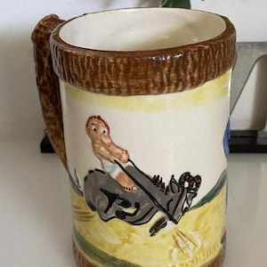 May include: A brown ceramic mug with a tree trunk handle. The mug features a hand-painted scene of a person riding a donkey. The mug has the text "Elgin, Ill." on the bottom.