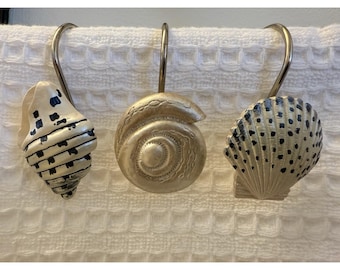 Puede incluir: Tres ganchos de cortina de ducha con forma de concha. Uno es una caracola beige con detalles azules, el segundo es una concha en espiral y el tercero es una concha de vieira con puntos azules. Todos tienen ganchos de metal plateado.