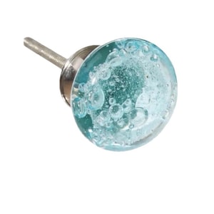 Puede incluir: Un pomo decorativo para armario con una cara de cristal transparente, redonda y azul claro. El cristal está lleno de pequeñas burbujas transparentes y está unido a una base y un tornillo de metal plateado.