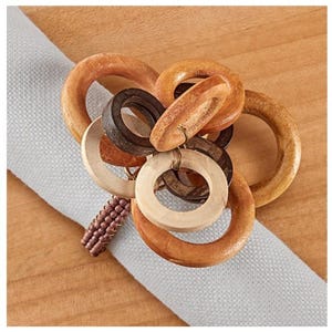 May include: A collection of wooden napkin rings in various shades of brown and natural wood tones. The rings are arranged in a cluster, with a beaded accent. The napkin rings are resting on a light gray napkin.