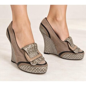 May include: A pair of taupe wedge shoes featuring a textured snakeskin-patterned panel on the toe and heel. The shoes include a decorative folded flap on the vamp with matching snakeskin trim and a thin black piping detail along the sides.