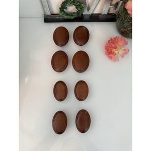 Può includere: Otto bottoni ovali in legno marrone sono disposti su una superficie bianca. I bottoni hanno un aspetto liscio e leggermente strutturato. I bottoni sono di un tono caldo e terroso e sono uniformemente distanziati.