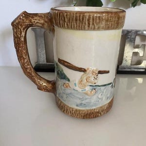 May include: A ceramic mug with a brown tree trunk handle and a white body. The mug features a painted scene of a bear in a forest setting.