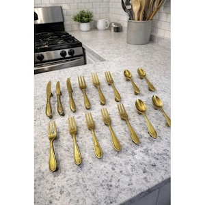 May include: A set of antique-style gold-coloured cutlery, including knives, forks, and spoons, arranged on a speckled countertop. The utensils have ornate handles and are displayed in a kitchen setting, suggesting a vintage or classic dining theme.