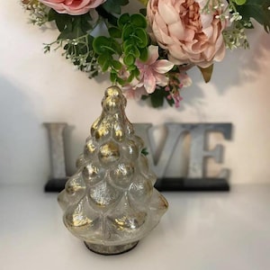Puede incluir: Un pequeño árbol de Navidad de cerámica blanca con detalles dorados. El árbol está decorado con un diseño texturizado y burbujeante.