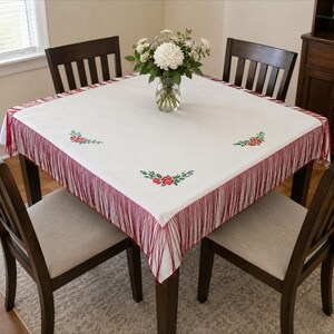 May include: A white tablecloth with red fringe and embroidered floral accents. The tablecloth is on a dark wood table, surrounded by four wooden chairs with beige seats. A vase of white flowers sits in the center.
