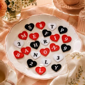 Puede incluir: Una colección de parches bordados en forma de corazón dispuestos sobre un plato blanco de borde festoneado. Los parches son rojos, blancos o negros, cada uno con una letra o número bordado en color contrastante, incluyendo S, 8, T, P, M, E, F, I, 9, 2, R, H, A, N, 0, 3, L y Y.