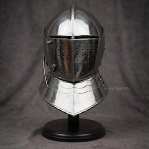May include: A polished silver-colored metal knight helmet featuring a rounded dome, a visor with a horizontal eye slit, and a faceplate with multiple ventilation holes. The helmet rests on a black circular stand and includes a neck guard.