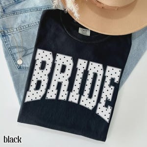 May include: A black short-sleeved t-shirt featuring the word BRIDE in large white block letters with a black polka dot pattern. The shirt has a Comfort Colors label at the inner neckline and is layered with light blue denim fabric.