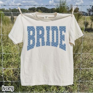 May include: An ivory short-sleeve t-shirt featuring the word BRIDE printed in a large, arched, blue collegiate-style font with a white polka dot pattern. The shirt is made of a solid, light-colored fabric and has a standard crew neckline.