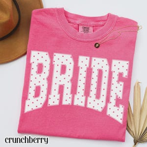 May include: A bright pink short-sleeve t-shirt featuring the word BRIDE in large white block letters with a pink polka dot pattern. The shirt has a Comfort Colors label at the inner neckline and is displayed with a brown hat and gold necklace.