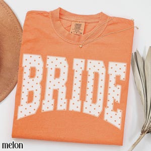 May include: A melon-colored short-sleeve t-shirt featuring the word BRIDE in large white block letters with a polka dot pattern. The shirt has a crew neckline and a Comfort Colors brand label printed on the inside of the collar.