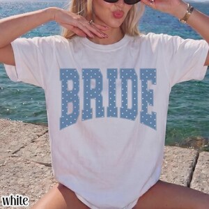 May include: A white short-sleeved t-shirt featuring the word BRIDE printed in large, block-style letters. The lettering is a dusty blue color and is decorated with a repeating pattern of small, white polka dots across the entire text.