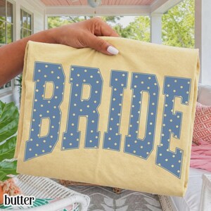 May include: A pale yellow t-shirt featuring the word BRIDE in large, block-style letters. The letters are blue with a repeating pattern of small, light yellow polka dots throughout the text, creating a bold and graphic design on the fabric.