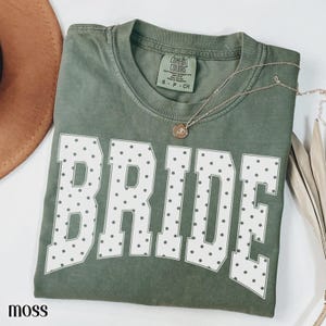May include: A moss green short-sleeved t-shirt featuring the word BRIDE in large white block letters with a dark polka dot pattern. The shirt has a crew neckline and a Comfort Colors brand label at the inner collar. A thin gold chain necklace rests on the fabric.