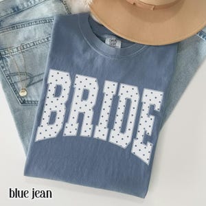 May include: A blue short-sleeved t-shirt featuring the word BRIDE in large white block letters with a dark polka dot pattern. The shirt has a Comfort Colors label at the inner neckline and is layered over light blue denim jeans.