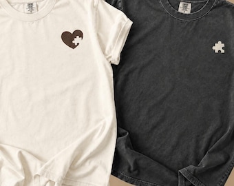 May include: A white t-shirt featuring a brown heart with a missing puzzle piece cutout on the left chest, paired with a charcoal gray t-shirt featuring a single white puzzle piece graphic on the left chest. Both shirts have crew necklines.