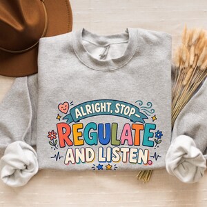 May include: A light grey crewneck sweatshirt featuring a colourful graphic print that reads "ALRIGHT, STOP REGULATE AND LISTEN" in bold, multi-coloured lettering, accented with small illustrations of a heart, flowers, stars, and wavy lines.