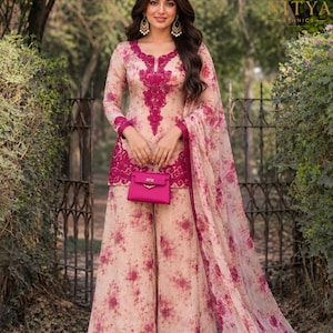 May include: A beige long-sleeved tunic and wide-leg pants set featuring a pink floral print, a dark pink embroidered neckline and hem, and a matching sheer floral dupatta. A small, solid pink handbag is held in front of the tunic.