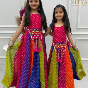 Pode incluir: Dois vestidos sem mangas com um padrão vibrante de riscas verticais multicoloridas e estampado de pontos brancos. Cada vestido inclui um painel triangular central adornado com espelhos refletores e borlas cor-de-rosa na cintura.