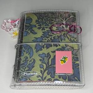 May include: A clear plastic binder featuring a green and blue floral patterned cover with the text Planwith-. It includes a pink rectangular insert with a lemon graphic, a metallic pink paperclip, and a star-shaped charm with a white flower.