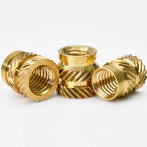 May include: Three cylindrical brass threaded inserts featuring a metallic gold finish. Each component displays a hollow center with internal screw threads and an exterior surface characterized by a distinct diagonal knurled pattern for grip.
