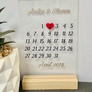 May include: A clear acrylic rectangular calendar sign featuring the names Aziliz & Steven in gold script, with black printed dates for August 2018. A red heart icon marks the number 2. The sign is held upright by a light-coloured wooden base.