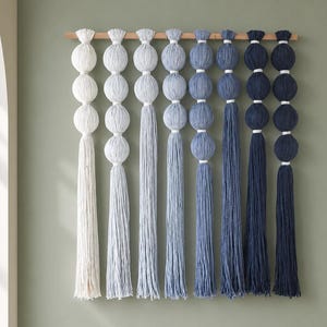 May include: A wall hanging featuring eight vertical strands of yarn suspended from a horizontal wooden rod. Each strand consists of stacked, spherical yarn balls in a gradient of blue shades, ranging from off-white to deep navy, ending in tassels.