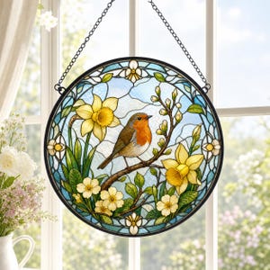 Könnte beinhalten: Ein runder Buntglas-Sonnenfänger mit einem Rotkehlchen auf einem Ast, umgeben von gelben Narzissen und kleinen weißen Blüten. Das Motiv ist in einen schwarzen Metallrahmen gefasst und verfügt über eine Metallkette zum Aufhängen.