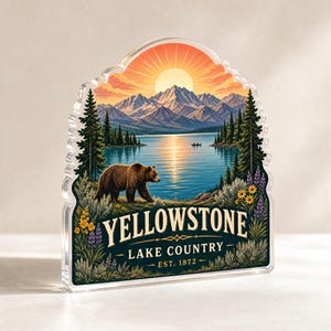 May include: A clear acrylic plaque featuring a colorful illustration of a brown bear walking near a lake with mountains and a sunrise in the background. The text reads YELLOWSTONE LAKE COUNTRY EST. 1872, surrounded by pine trees and wildflowers.