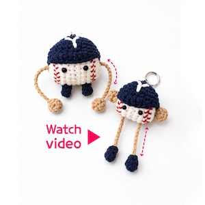 May include: Two crocheted baseball keychains with dark blue caps, white bodies with red stitching, black bead eyes, and tan limbs. One figure has arms extended, the other has long legs. Text reads Watch video with a pink play button icon.