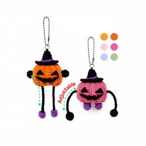 Op de afbeelding: Twee gehaakte pompoen-sleutelhangers met zwarte heksenhoeden en jack-o'-lantern gezichten. De ene is oranje en de andere roze, beide met zwarte ledematen en paarse voeten. Het woord Adjustable staat in het rood met groene pijlen voor beweging.