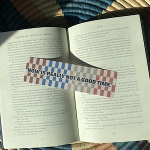 May include: A rectangular bookmark with a checkered pattern in shades of blue, white, and red. The text "NOW IS REALLY NOT A GOOD TIME." is printed in black capital letters across the center of the bookmark, which rests on an open book.