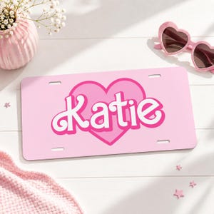 May include: A rectangular light pink license plate featuring a large dark pink heart graphic in the center. The name Katie is printed across the heart in a bold white font with a dark pink outline. The plate includes four mounting slots.