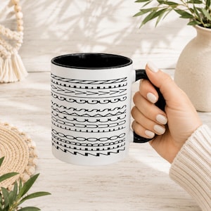 May include: A white ceramic mug with a black interior and handle, featuring a series of horizontal black geometric patterns and the text "ZERO FUCKS GIVEN" integrated into the design. The mug has a cylindrical shape and a smooth finish.